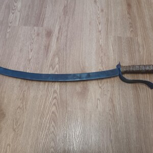 May include: A curved, dark metal sword with a textured, wrapped handle. The blade has a slight curve and a pointed tip. The handle is brown and the guard is black. The sword is lying on a wooden surface.