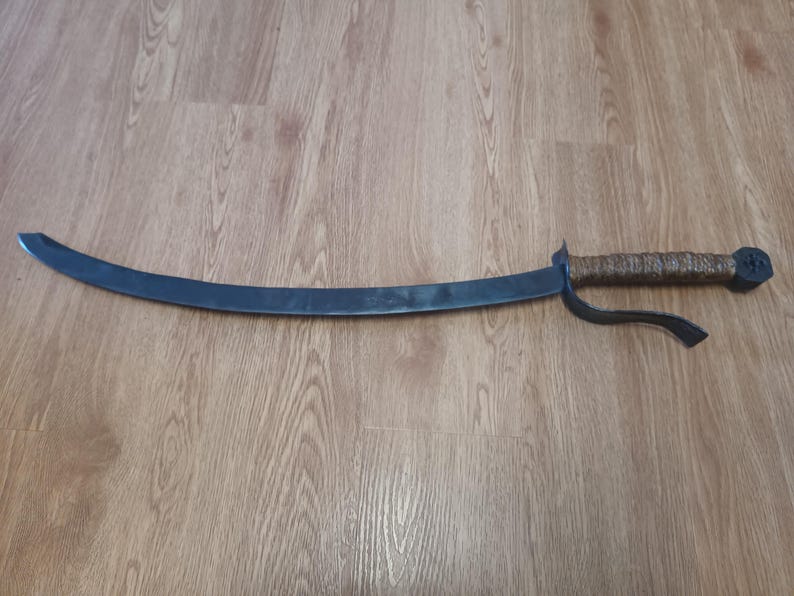 May include: A curved, dark metal sword with a textured, wrapped handle. The blade has a slight curve and a pointed tip. The handle is brown and the guard is black. The sword is lying on a wooden surface.
