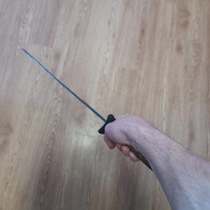 May include: A black-handled sword with a long, thin, silver blade. The sword is held in a hand against a light brown wooden background. The handle is black and the blade is straight.