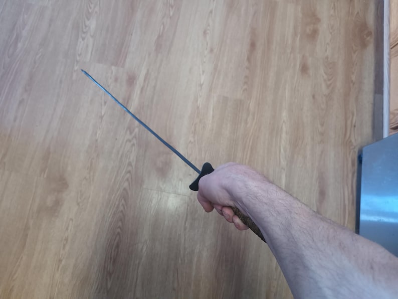 May include: A black-handled sword with a long, thin, silver blade. The sword is held in a hand against a light brown wooden background. The handle is black and the blade is straight.