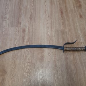 May include: A curved, dark metal sword with a wrapped handle and a distinctive guard. The blade has a sharp point, and the handle is wrapped in a textured material. The sword is lying on a wooden surface.