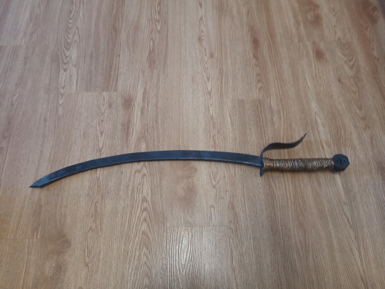 May include: A curved, dark metal sword with a wrapped handle and a distinctive guard. The blade has a sharp point, and the handle is wrapped in a textured material. The sword is lying on a wooden surface.