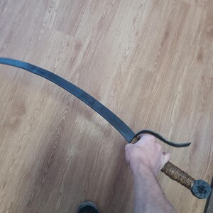 May include: A curved, black metal sword with a textured, wrapped handle. The blade has a slight curve and a pointed tip. The handle is wrapped in a brown material and has a decorative end.