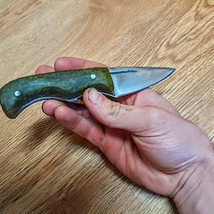 Pode incluir: Uma pequena faca com cabo verde e lâmina prateada. A faca está sendo segurada na mão, com a lâmina apontando para a direita.