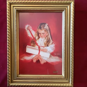 May include: A framed print of a young child opening a gift. The child is wearing a white dress with red accents and a white bow in their hair. The frame is gold with a textured edge. The background is red.