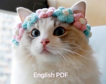 Puede incluir: Un gato blanco con ojos azules que lleva una corona de flores de ganchillo con flores rosas y azules.