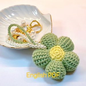 May include: A crocheted flower charm in green and yellow. The flower has six petals and a yellow centre. The charm is hanging from a string of pearls.