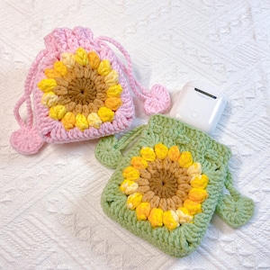 Könnte beinhalten: Zwei gehäkelte Sonnenblumenbeutel, einer rosa und einer grün, mit einem weißen AirPod-Etui im grünen Beutel.