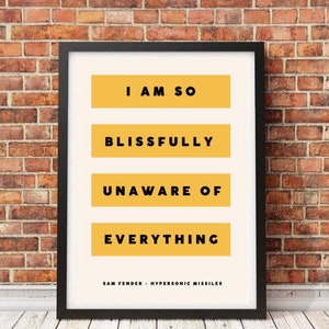 May include: A black framed print with a white background and yellow text. The text reads "I am so blissfully unaware of everything" and "Sam Fender - Hypersonic Missiles".
