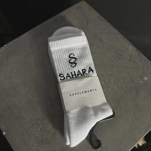 May include: White socks featuring the word "SAHARA" in black, with a snake graphic. The socks have a tag that reads "SUPPLEMENTS". The socks are displayed on a grey surface.