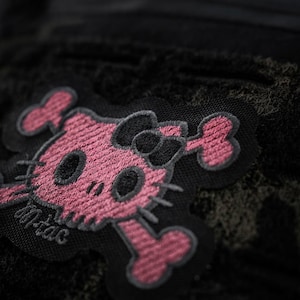 May include: A black embroidered patch featuring a pink skull design with cat-like ears, a bow, and whiskers, arranged in a crossbones style. The text M-TAC is stitched in grey thread on the lower left side of the patch, which is set against a dark fabric.