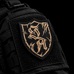 Puede incluir: Un parche táctico negro con un diseño en forma de escudo y letras estilizadas en relieve de color beige. El parche está sujeto a una superficie de tela negra con correas integradas y un panel de cierre de gancho y bucle.