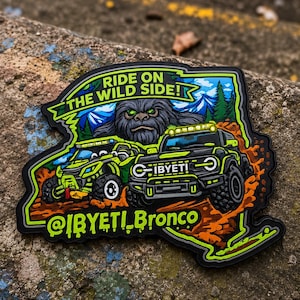 May include: A die-cut patch featuring a graphic illustration of a green off-road vehicle and a black SUV driving through mud. A large yeti figure is in the background with mountains and trees. The text reads RIDE ON THE WILD SIDE! and @IBYETI_Bronco.