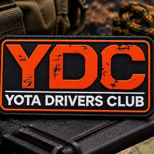 May include: A rectangular black patch with an orange border featuring the large orange letters YDC with a distressed texture. Below, a thin white horizontal line separates the text YOTA DRIVERS CLUB in white capital letters on a black background.