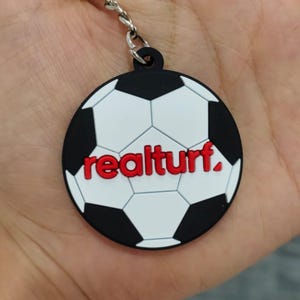May include: A circular keyring featuring a black and white football pattern with the word "realturf." printed in bold red letters across the centre. The keyring is attached to a silver metal split ring by a short silver metal chain.