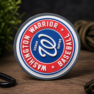Puede incluir: Un parche circular de goma con un anillo exterior rojo que contiene el texto blanco WASHINGTON WARRIOR BASEBALL y dos estrellas blancas. El centro es un círculo azul con un logotipo de W cursiva blanca y el texto CEDAR RAPIDS en letras pequeñas.