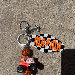 May include: A keychain with a black and white checkered tag that reads "MAZMA" in orange letters. The keychain also features a small race car charm and a decorative orange bead.
