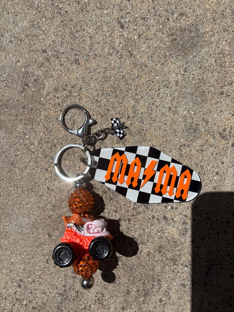 May include: A keychain with a black and white checkered tag that reads "MAZMA" in orange letters. The keychain also features a small race car charm and a decorative orange bead.