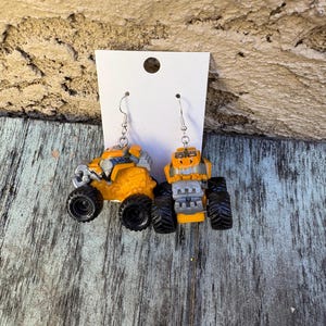K&ouml;nnte beinhalten: Ein Paar ausgefallene Ohrringe in Form von Miniatur-Monster Trucks. Die Ohrringe sind haupts&auml;chlich gelb mit schwarzen Reifen und silberfarbenen Haken. Die Trucks haben detaillierte Merkmale, einschlie&szlig;lich K&uuml;hlergrills und &Uuml;berrollk&auml;figen.