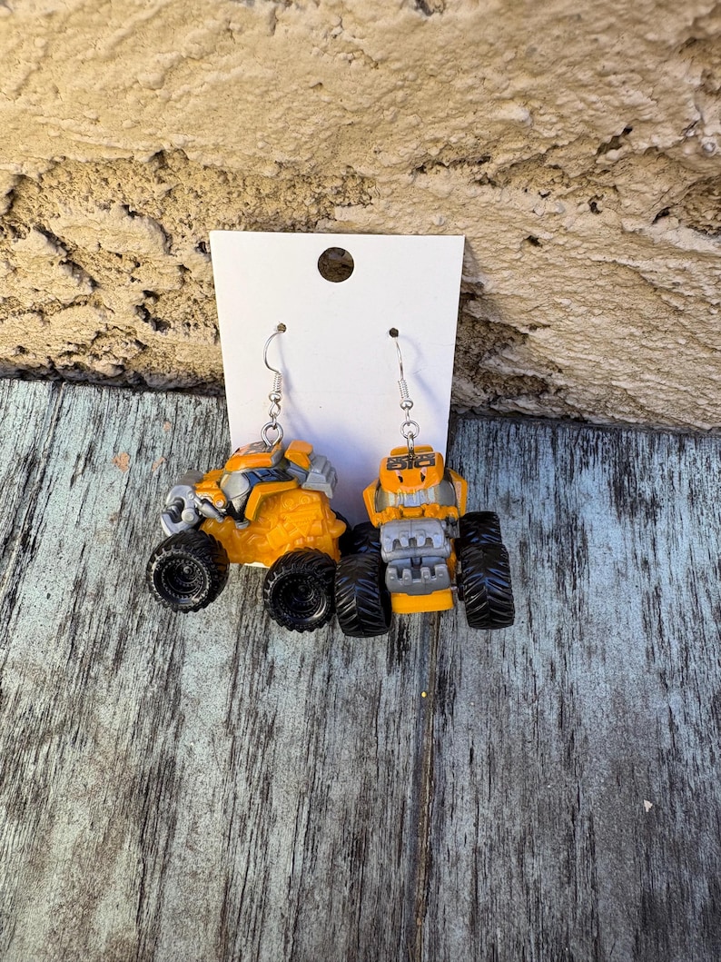 K&ouml;nnte beinhalten: Ein Paar ausgefallene Ohrringe in Form von Miniatur-Monster Trucks. Die Ohrringe sind haupts&auml;chlich gelb mit schwarzen Reifen und silberfarbenen Haken. Die Trucks haben detaillierte Merkmale, einschlie&szlig;lich K&uuml;hlergrills und &Uuml;berrollk&auml;figen.