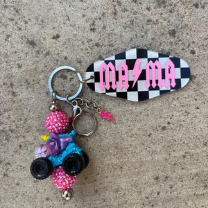 May include: A keychain featuring a blue monster truck with a unicorn head, black wheels, and pink beaded accents. The keychain includes a tag with the word "MAMA" in pink gothic lettering over a black and white checkered background.