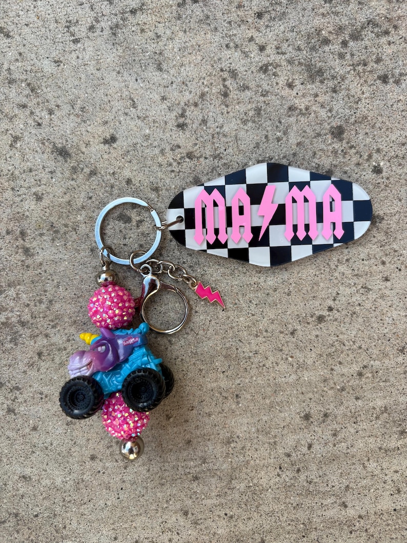 May include: A keychain featuring a blue monster truck with a unicorn head, black wheels, and pink beaded accents. The keychain includes a tag with the word "MAMA" in pink gothic lettering over a black and white checkered background.