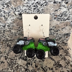 Puede incluir: Un par de pendientes novedosos con forma de camiones monstruo en miniatura. Los camiones son negros y verdes con ganchos plateados. El texto "Grave Digger" es visible en la parte superior de cada cami&oacute;n.