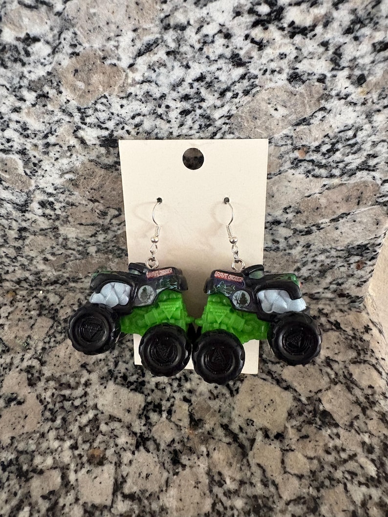 Puede incluir: Un par de pendientes novedosos con forma de camiones monstruo en miniatura. Los camiones son negros y verdes con ganchos plateados. El texto "Grave Digger" es visible en la parte superior de cada cami&oacute;n.