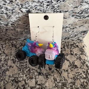 Peut inclure: Boucles d'oreilles fantaisie avec des licornes miniatures en forme de monster truck. Les boucles d'oreilles pr&eacute;sentent des t&ecirc;tes de licorne bleues, violettes et roses avec des cornes jaunes, mont&eacute;es sur des roues noires. Elles sont suspendues &agrave; des crochets d'oreilles argent&eacute;s.
