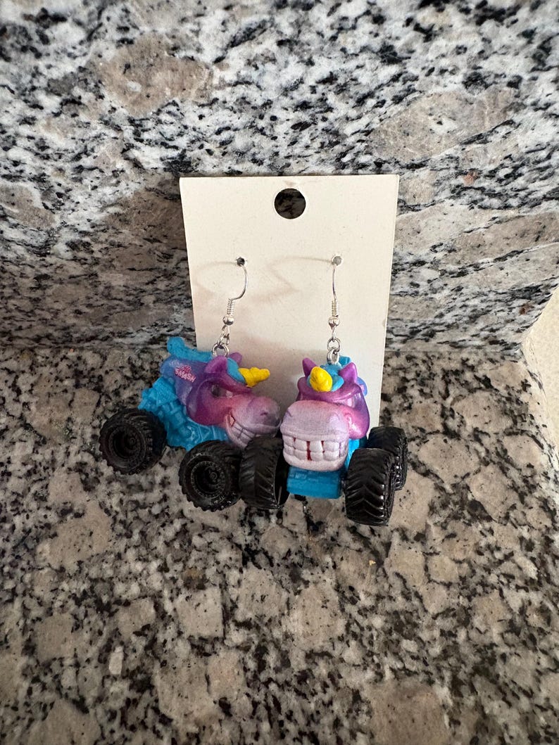 Peut inclure: Boucles d'oreilles fantaisie avec des licornes miniatures en forme de monster truck. Les boucles d'oreilles pr&eacute;sentent des t&ecirc;tes de licorne bleues, violettes et roses avec des cornes jaunes, mont&eacute;es sur des roues noires. Elles sont suspendues &agrave; des crochets d'oreilles argent&eacute;s.