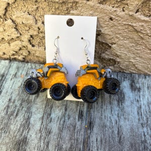 K&ouml;nnte beinhalten: Ein Paar ausgefallene Ohrringe mit Miniatur-Monster-Trucks in Orange und Grau mit schwarzen Reifen. Die Ohrringe sind an silberfarbenen Fischhaken-Ohrdr&auml;hten befestigt und werden auf einer wei&szlig;en Karte pr&auml;sentiert.