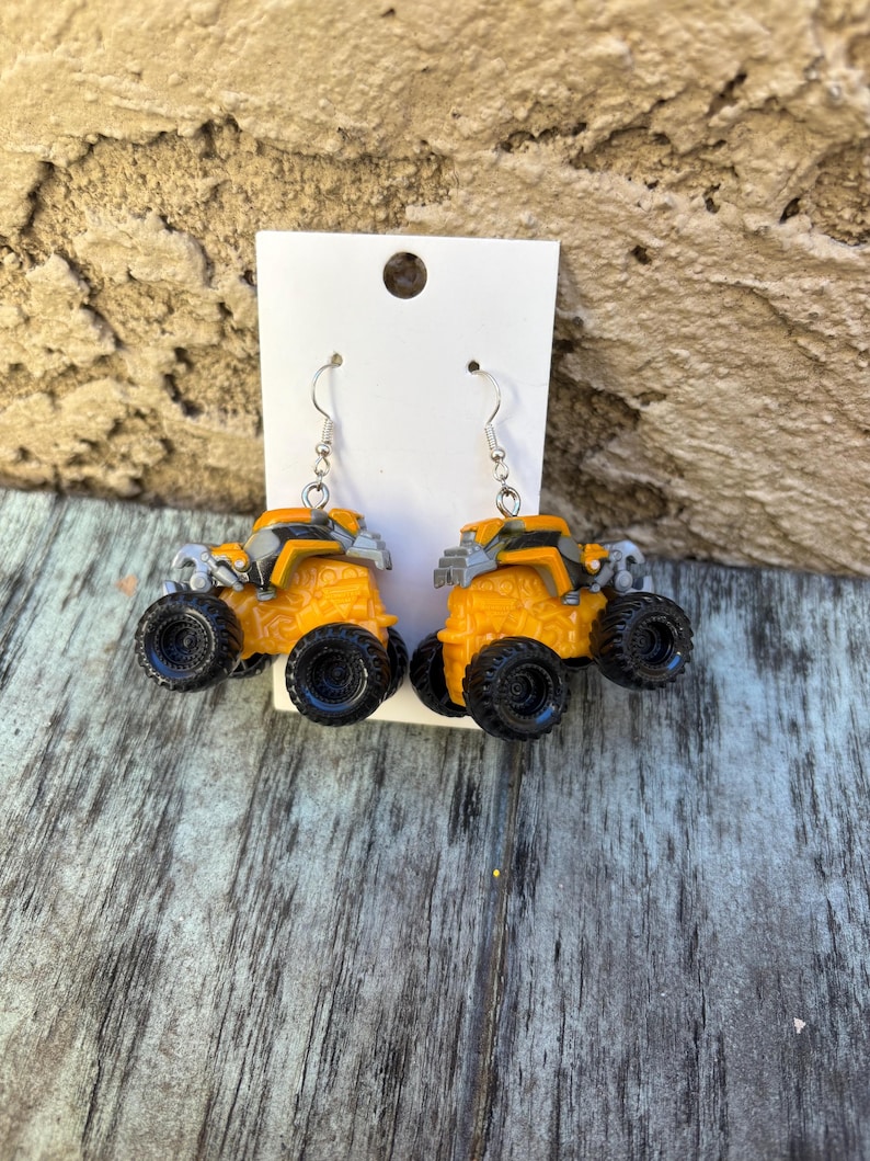 K&ouml;nnte beinhalten: Ein Paar ausgefallene Ohrringe mit Miniatur-Monster-Trucks in Orange und Grau mit schwarzen Reifen. Die Ohrringe sind an silberfarbenen Fischhaken-Ohrdr&auml;hten befestigt und werden auf einer wei&szlig;en Karte pr&auml;sentiert.