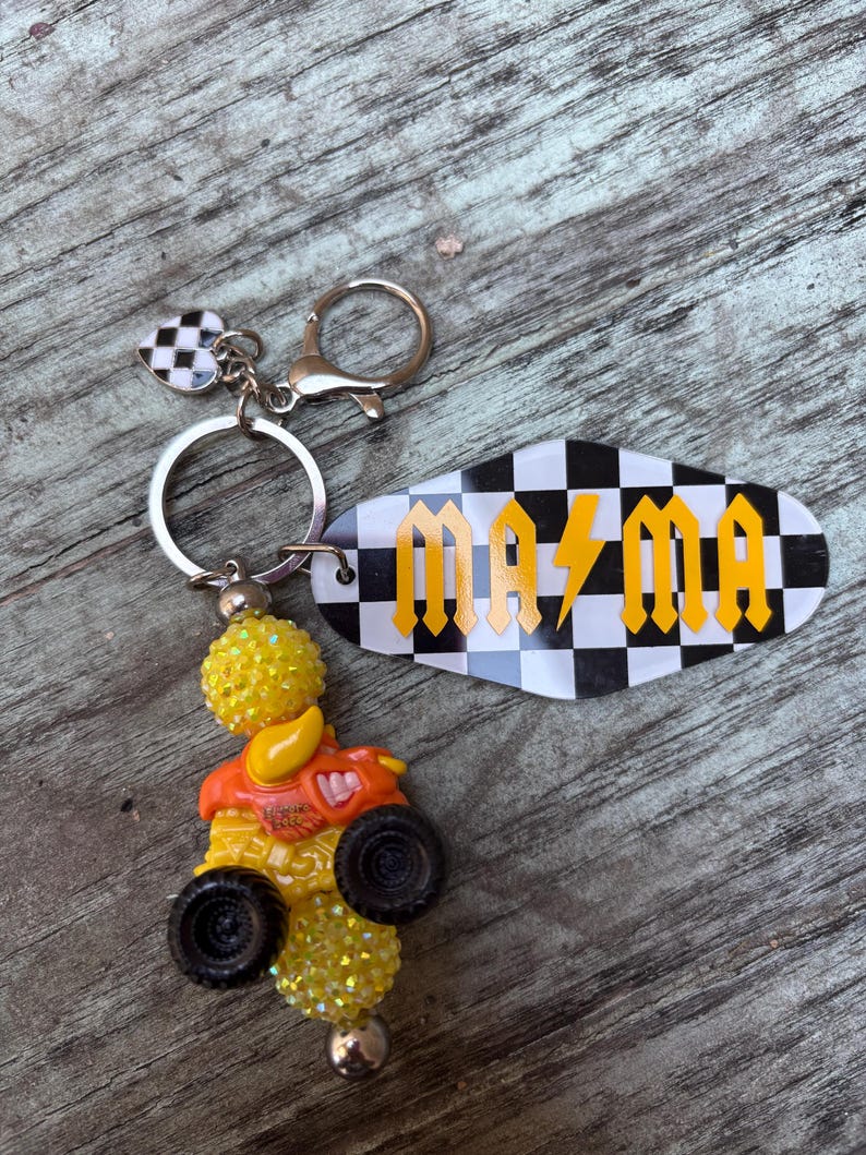 May include: A keychain with a black and white checkered heart charm and a key tag that reads "MAMA" in yellow letters. The keychain also features a yellow and orange monster truck charm with black wheels.