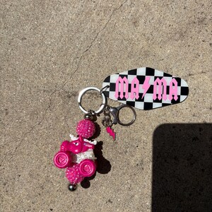 May include: A pink keychain featuring a small ATV with glitter accents and a decorative charm. The keychain includes a black and white checkered tag with the word "MAZMA" in pink gothic lettering and a pink lightning bolt charm.