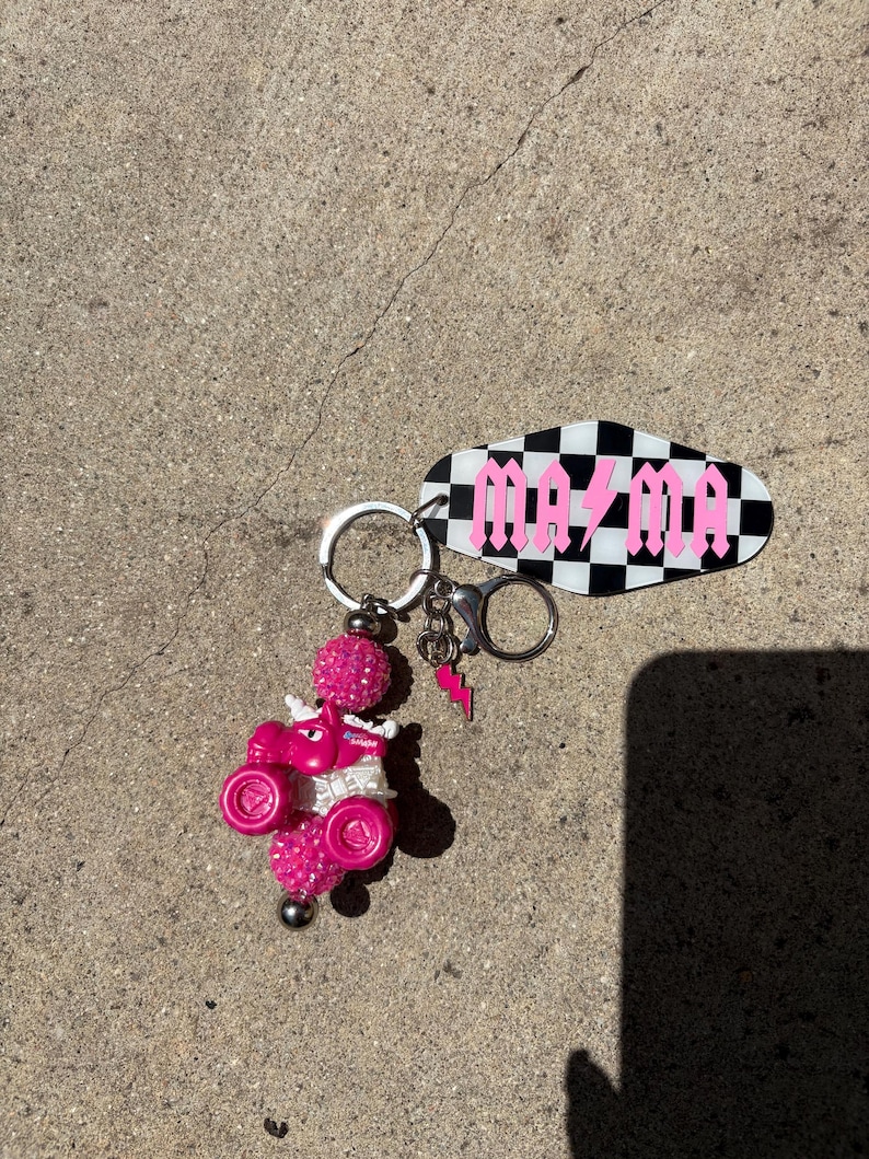 May include: A pink keychain featuring a small ATV with glitter accents and a decorative charm. The keychain includes a black and white checkered tag with the word "MAZMA" in pink gothic lettering and a pink lightning bolt charm.