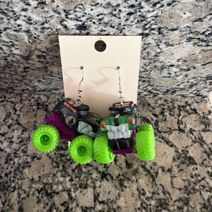 May include: A pair of monster truck earrings. One earring is black and purple with green wheels, the other is green and black with green wheels. The earrings hang from silver-coloured fish hook ear wires.