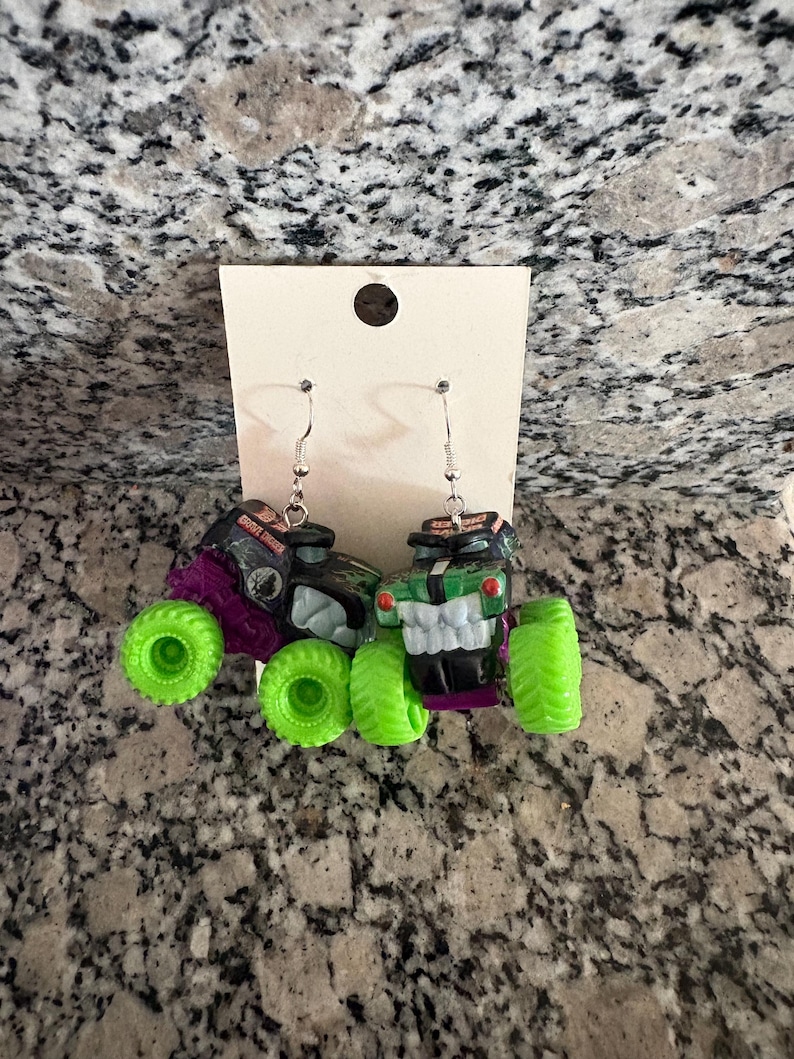 May include: A pair of monster truck earrings. One earring is black and purple with green wheels, the other is green and black with green wheels. The earrings hang from silver-coloured fish hook ear wires.