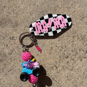 May include: A keychain with a black and white checkered oval tag that reads "MA MA" in pink block letters. The keychain has a small pink lightning bolt charm and a miniature monster truck with a unicorn on top.