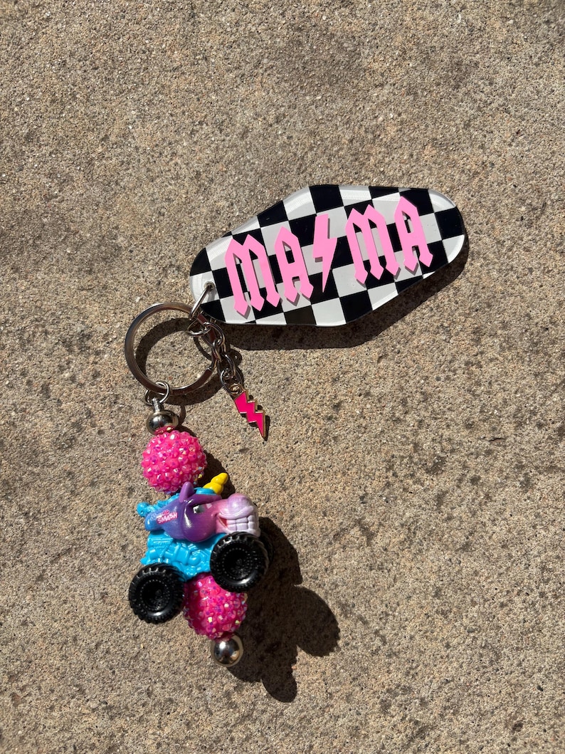 May include: A keychain with a black and white checkered oval tag that reads "MA MA" in pink block letters. The keychain has a small pink lightning bolt charm and a miniature monster truck with a unicorn on top.