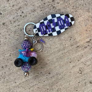 May include: A handmade keychain featuring a black and white checkered tag with the word "MAYA" in purple lettering. The keychain also includes a charm with a small blue monster truck, purple beads, and a lightning bolt charm.