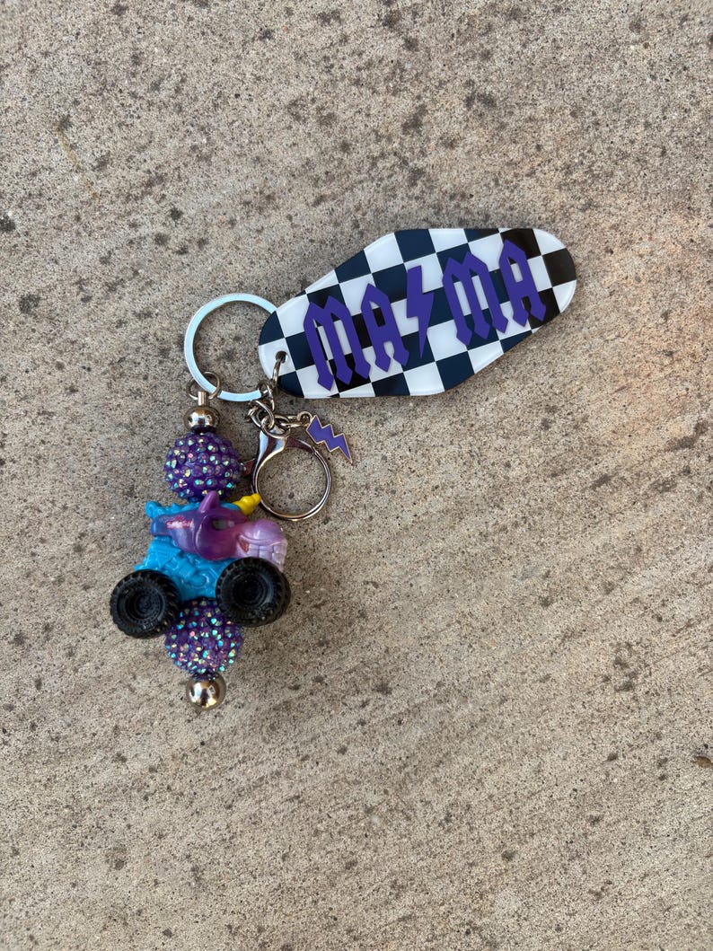 May include: A handmade keychain featuring a black and white checkered tag with the word "MAYA" in purple lettering. The keychain also includes a charm with a small blue monster truck, purple beads, and a lightning bolt charm.