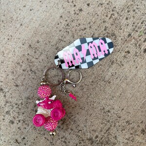 May include: A keychain with a black and white checkered tag that reads "mama" in pink gothic lettering. The keychain also features a pink and white beaded charm with a pink dragon and a pink lightning bolt charm.