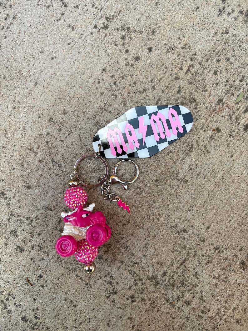 May include: A keychain with a black and white checkered tag that reads "mama" in pink gothic lettering. The keychain also features a pink and white beaded charm with a pink dragon and a pink lightning bolt charm.