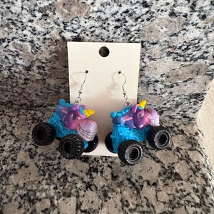 Peut inclure: Boucles d'oreilles fantaisie avec un design de monster truck sur le th&egrave;me de la licorne. Les boucles d'oreilles ont des roues noires, des corps bleus et violets et des cornes jaunes. Le mot "PASH" est visible sur le dessus du camion.