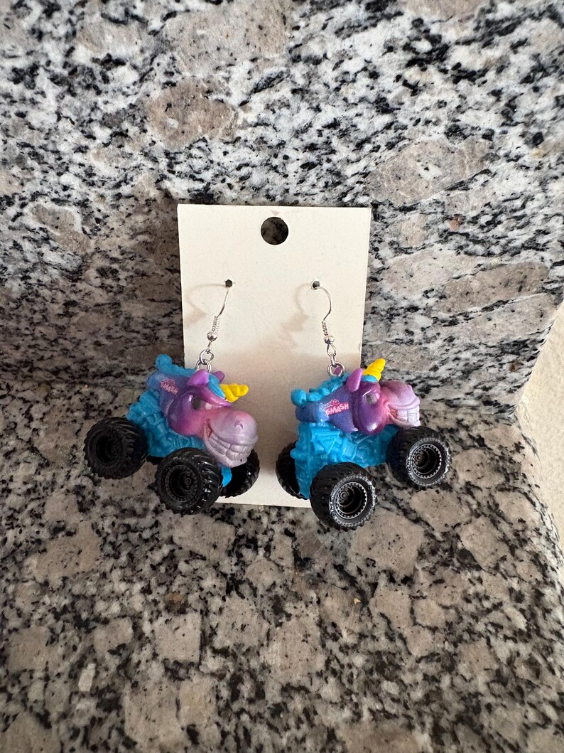 Peut inclure: Boucles d'oreilles fantaisie avec un design de monster truck sur le th&egrave;me de la licorne. Les boucles d'oreilles ont des roues noires, des corps bleus et violets et des cornes jaunes. Le mot "PASH" est visible sur le dessus du camion.