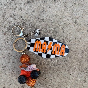May include: A keychain featuring a black and white checkered oval with the word "MAMA" in orange lettering. The keychain includes a small race car charm and a checkered flag charm. The beads are orange and black.