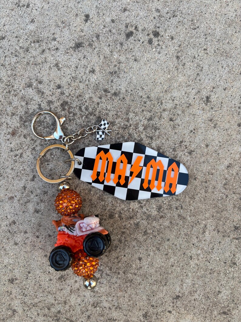 May include: A keychain featuring a black and white checkered oval with the word "MAMA" in orange lettering. The keychain includes a small race car charm and a checkered flag charm. The beads are orange and black.