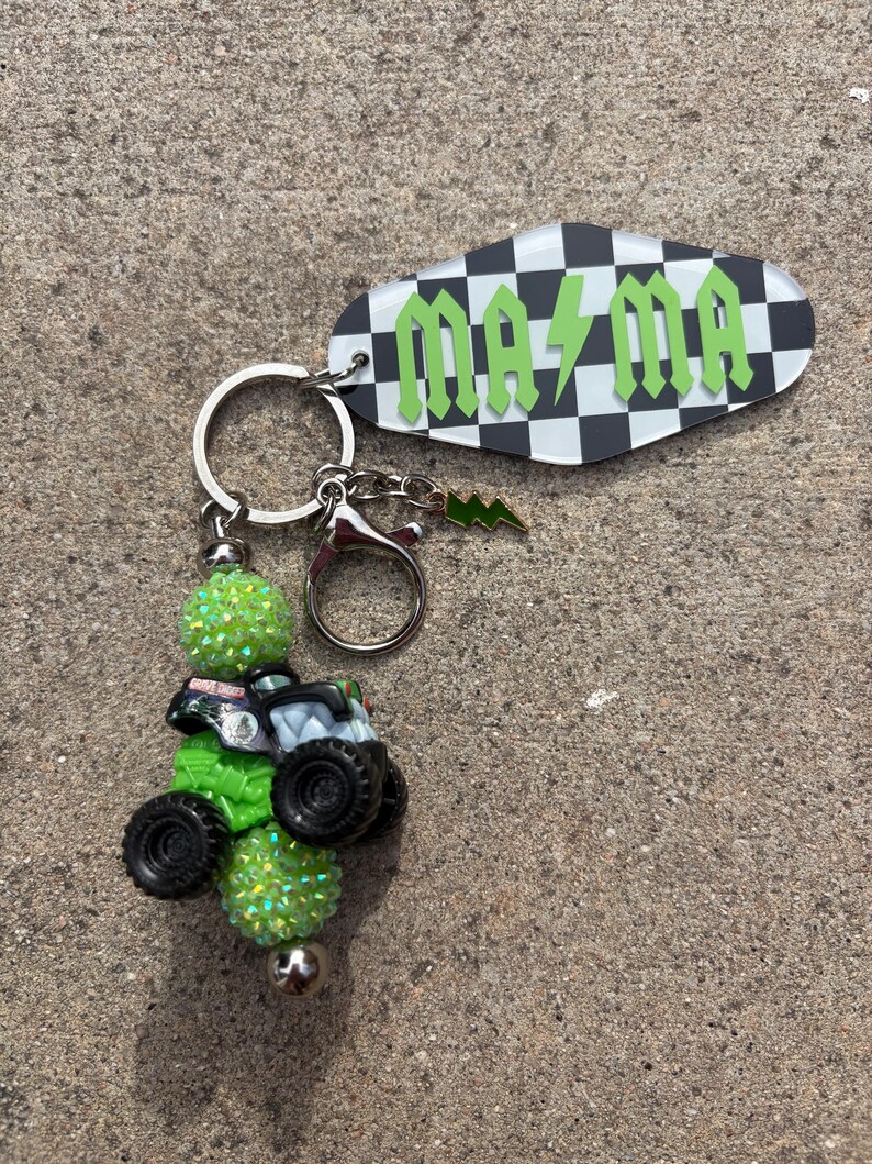 May include: Keychain with a green monster truck charm, green glitter beads, and a tag with the word "MAMA" in green lettering on a black and white chequered background. Includes a silver keyring and a lightning bolt charm.
