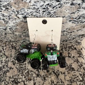 Puede incluir: Pendientes novedosos con dise&ntilde;os de monster trucks en miniatura. Un cami&oacute;n es negro y verde, el otro verde y blanco. Ambos tienen ruedas negras y ganchos plateados. Los pendientes se muestran en una tarjeta blanca.
