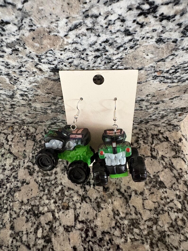 Puede incluir: Pendientes novedosos con dise&ntilde;os de monster trucks en miniatura. Un cami&oacute;n es negro y verde, el otro verde y blanco. Ambos tienen ruedas negras y ganchos plateados. Los pendientes se muestran en una tarjeta blanca.